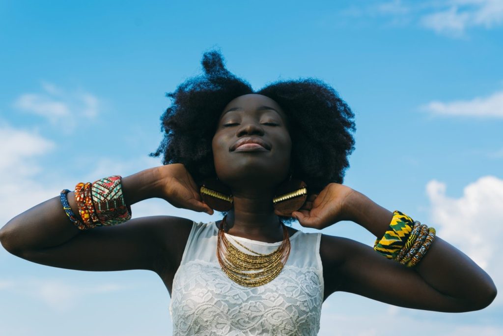 Color photo of a Black woman with her hands behind her head, eyes closed and her calm face turned up toward the sky.