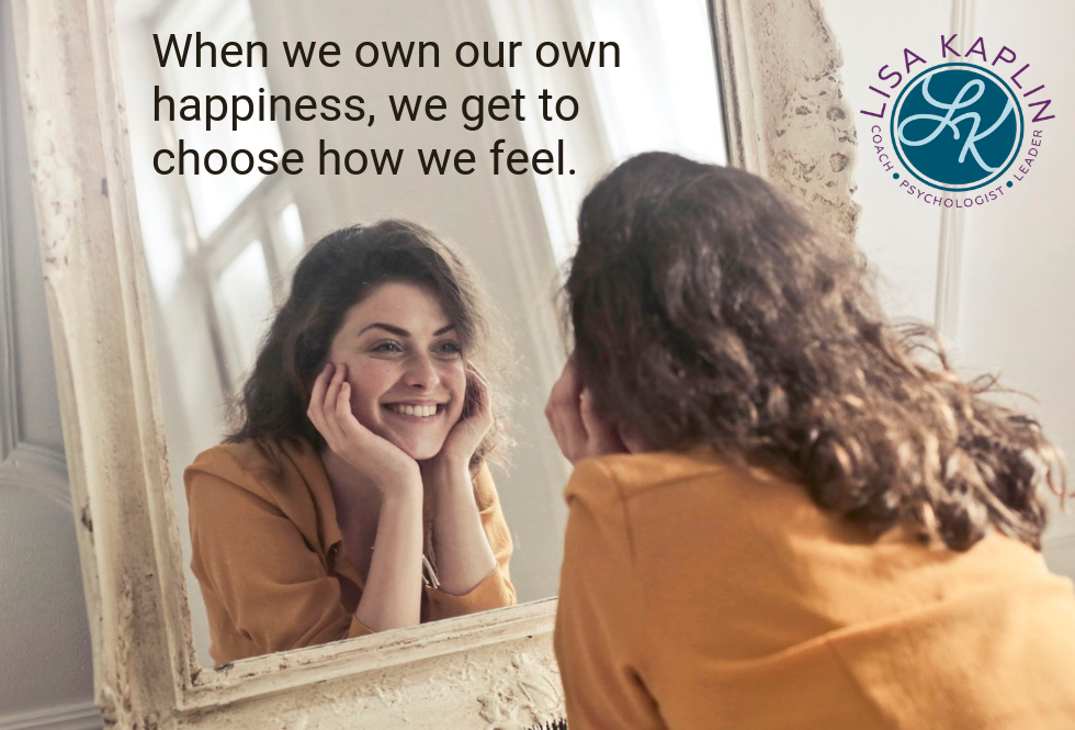 A photo of a young white woman staring into a mirror. We see her reflection and she is smiling. Above her reflection is the text “When we own our own happiness, we get to choose how we feel.” The Lisa Kaplin logo is in the top right corner.