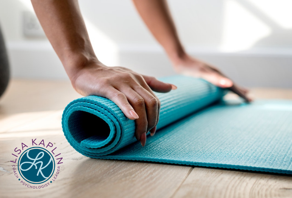 A closeup color photo of a white woman’s hands as she rolls up a turquoise blue yoga mat. The Lisa Kaplin logo is in the bottom left corner.