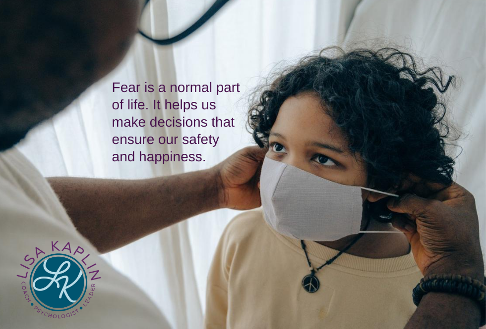 A color photo an elementary school age Black child looking up at their parent who is helping them put on a face mask. The text in the middle of the image reads “Fear is a normal part of life. It helps us make decisions that ensure our safety and happiness?” The Lisa Kaplin logo is in the bottom left corner.