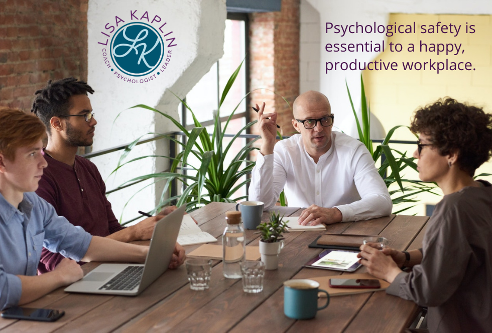 A color photo of a workplace meeting. A white, middle-age man in a white dress shirt sits at the head of the table. A younger white man and a young Black man are to his left. A middle-age white woman is to his right. All look tense. The text at the top right reads “Psychological safety is essential to a happy productive workplace” The Lisa Kaplin logo is in the top left corner.