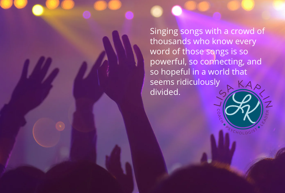 A color photo of a crowd at a concert taken from the back and in shadow. The tops of people’s heads and their raised open hands are visible against a background of purple stage lights. The text on the right reads “Singing songs with a crowd of thousands who know every word of those songs is so powerful, so connecting, and so hopeful in a world that seems ridiculously divided.” The Lisa Kaplin logo is beneath the text.