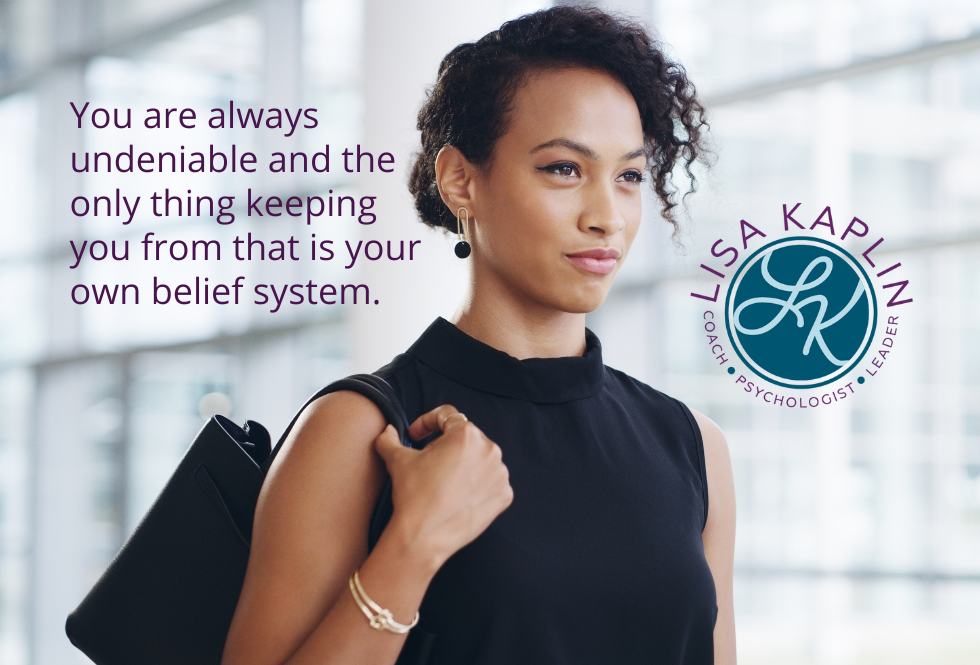 A color photo of a Black woman in black business attire holding a handbag on her shoulder. She is facing to the right and looking ahead with a determined expression. Behind her is an out of focus wall of windows. The text on her left reads “You are always undeniable and the only thing keeping you from that is your own belief system.“ The Lisa Kaplin logo is ion her right.